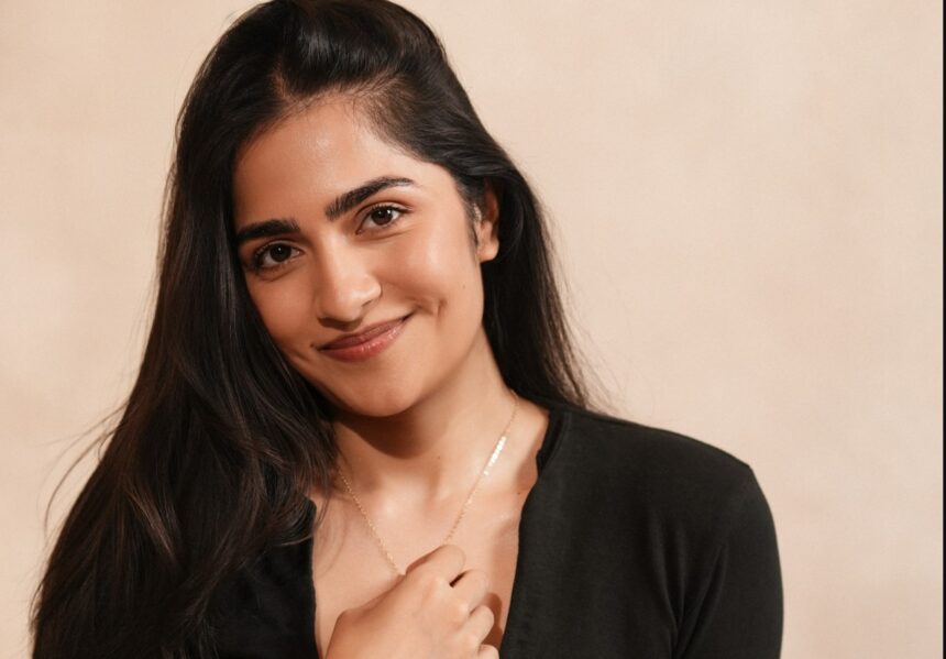 Rukmini Vasanth wearing a black top and relaxed light-wash jeans in a warm studio portrait